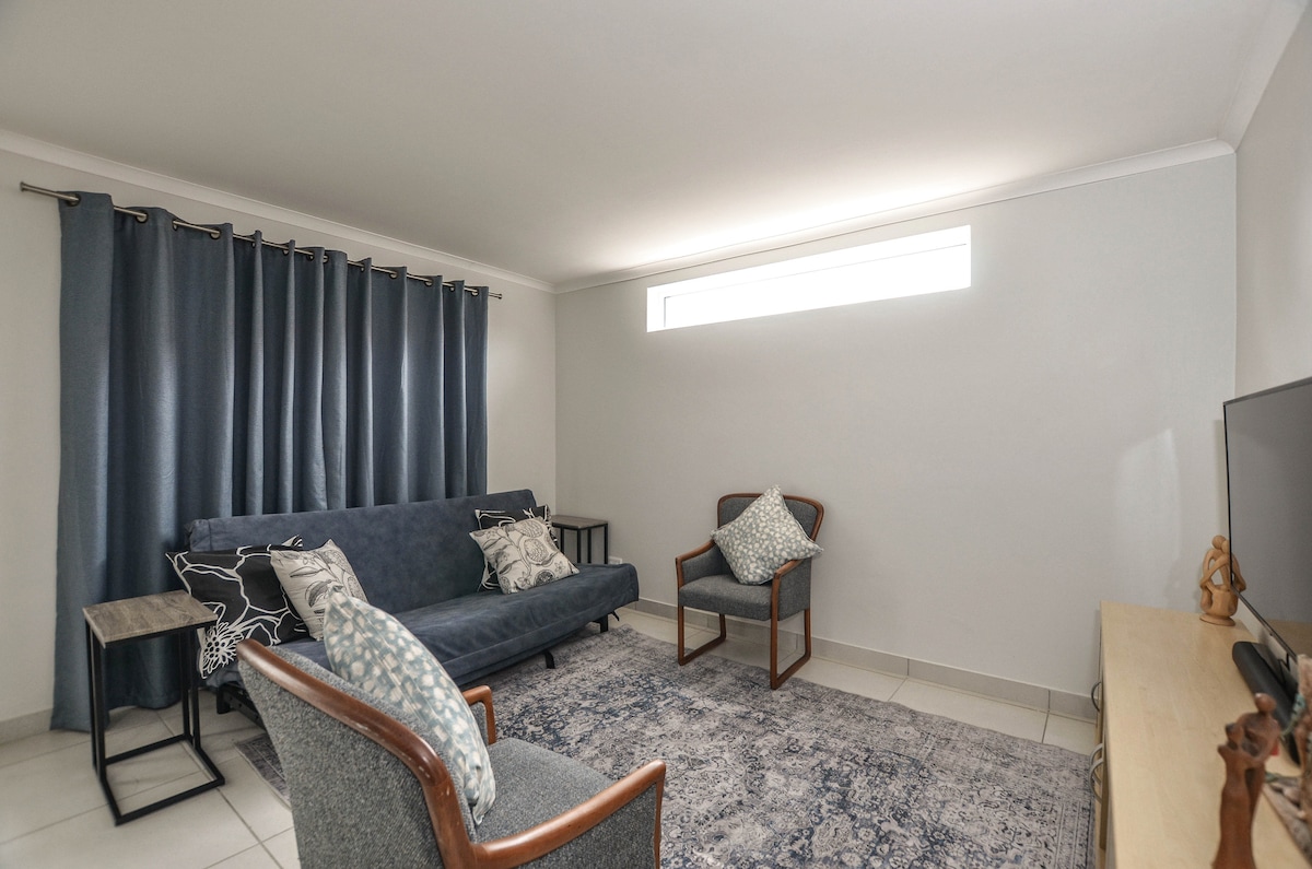 A cozy lounge area features a dark blue sofa with decorative pillows, accompanied by two armchairs. A small side table sits beside the sofa, while a light-colored rug covers the tiled floor. Natural light filters in through a high window, enhancing the space's welcoming feel.