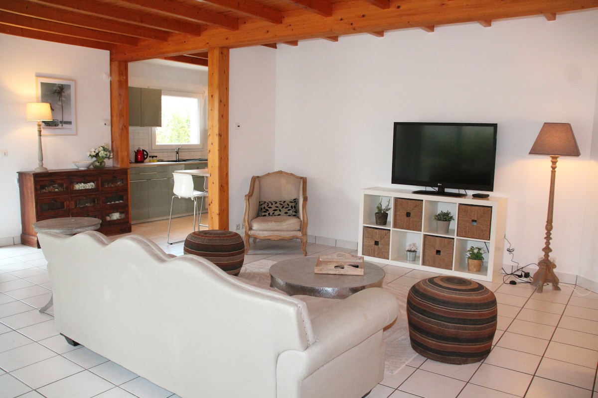 A spacious living area is presented with a comfortable couch and decorative poufs. A flat-screen television is mounted on the wall, while a wooden side table and lamp provide additional lighting. Open spaces lead to a modern kitchen area visible in the background.