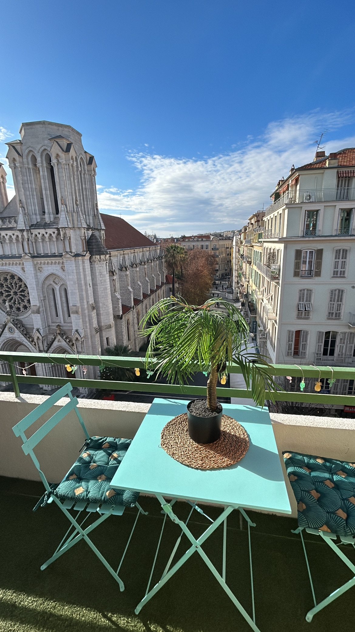 A small balcony features a round table and two folding chairs, both in teal. A potted palm adds greenery. In the background, the view includes a church with detailed architecture and a street that stretches into the distance under a clear blue sky.