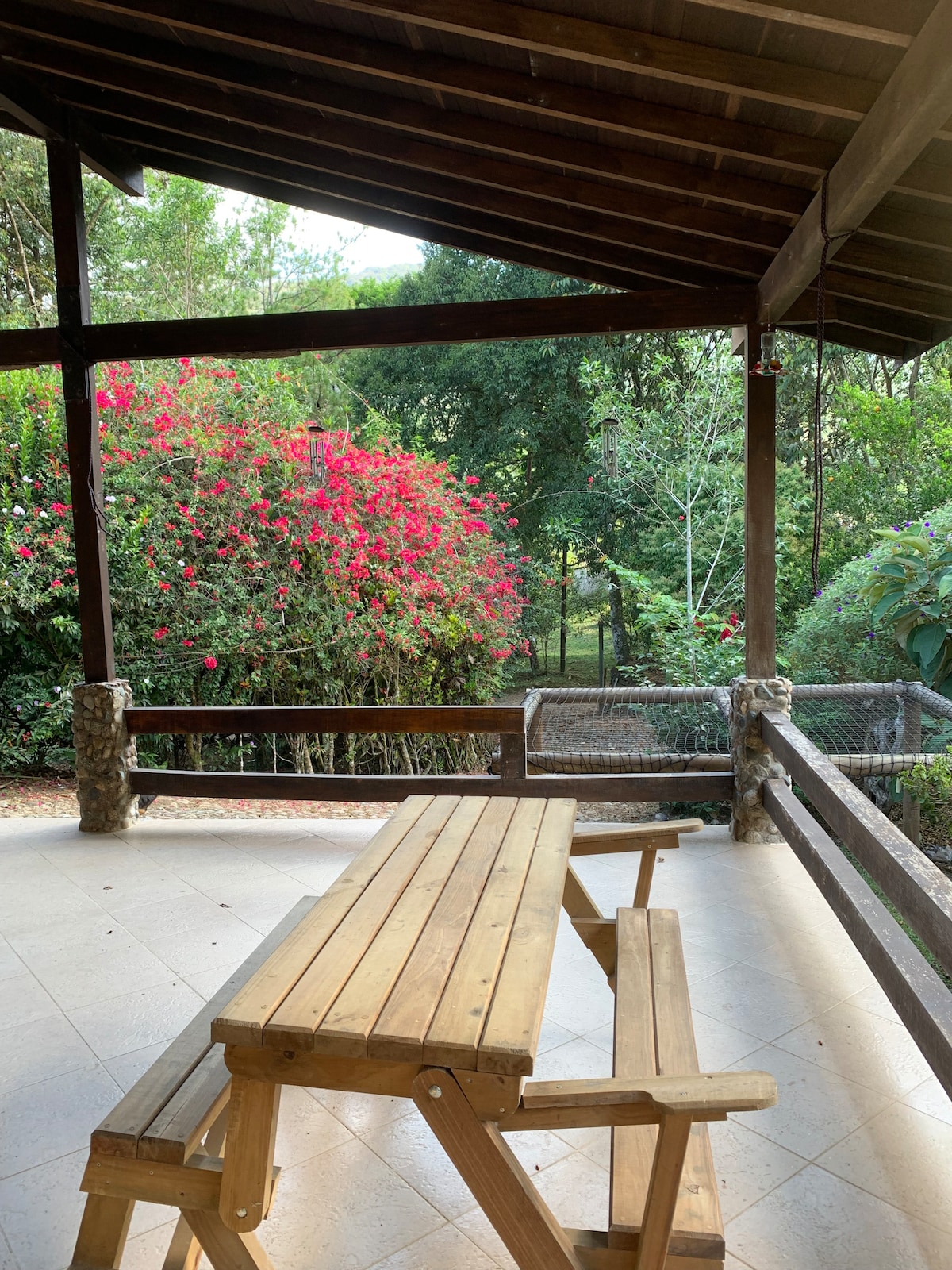A shaded outdoor space is featured with a wooden picnic table, surrounded by lush greenery and vibrant flowering plants. The area offers a peaceful view of the surrounding gardens and natural landscape, creating a serene setting for relaxation.
