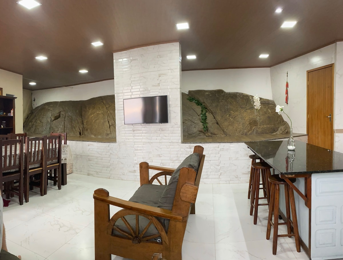 The spacious living area features a natural stone formation integrated into the design, with a flat-screen television mounted on a white tiled wall. Wooden dining tables and chairs sit nearby, complemented by dark wooden seating arrangements. The room is illuminated by recessed ceiling lights.
