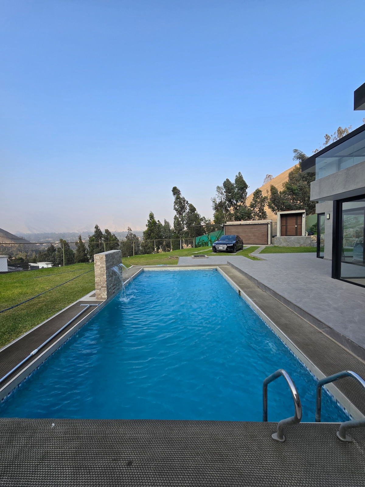 A serene swimming pool is presented, featuring clear blue water and modern steps for easy access. Lush greenery surrounds the area, with distant hills visible under a clear sky. A stylish home is located nearby, complemented by a parking area.