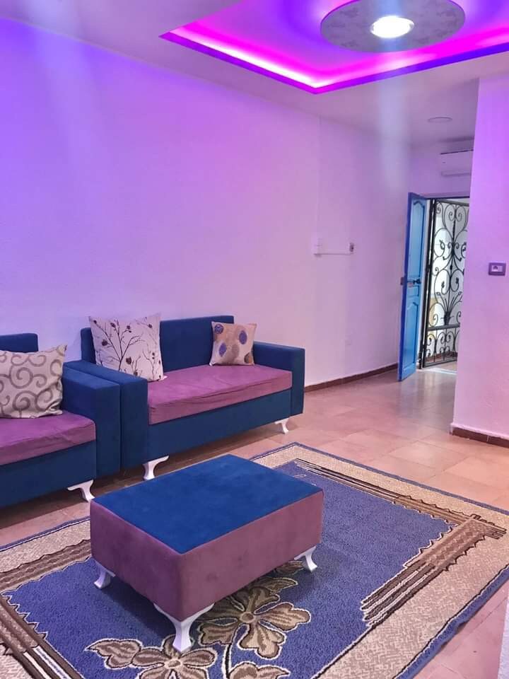 The living area features a combination of blue and purple furnishings, including a sofa set adorned with decorative pillows. A soft rug lies beneath a low table, and vibrant purple lighting enhances the modern ceiling design, creating a welcoming space.