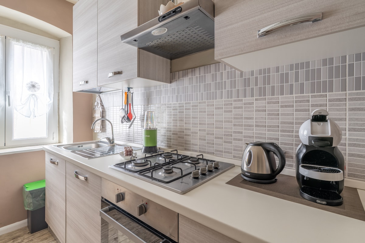 A modern kitchen features light wood cabinetry and a neutral tile backsplash. The space includes a gas stove, an electric kettle, and a coffee maker. Natural light filters in through a window adorned with a sheer curtain, with a small green trash can visible in the corner.