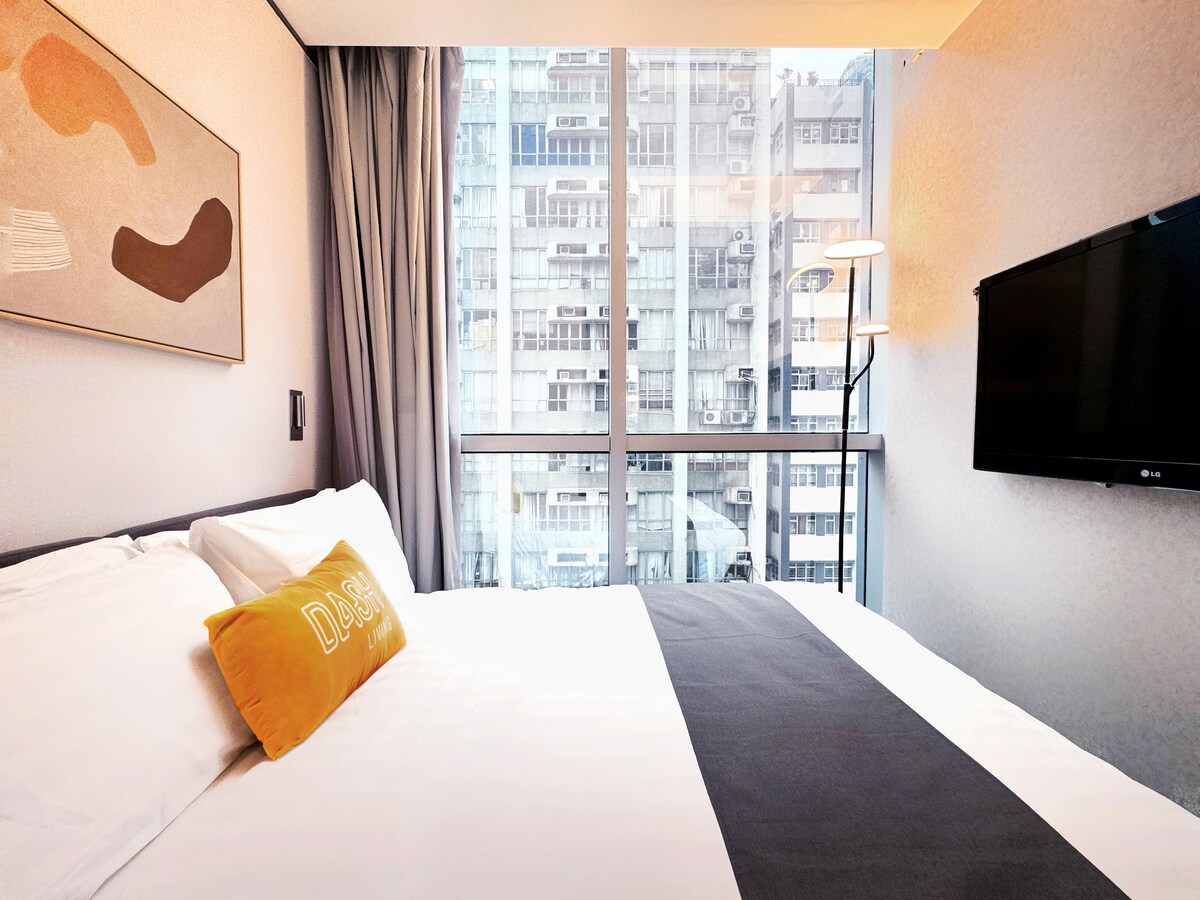 A spacious bed is framed by soft, neutral-toned walls, with light filtering in through large floor-to-ceiling windows. A modern artwork is displayed above the bed, and a flat-screen TV is mounted on the wall opposite, providing a functional and stylish ambiance.