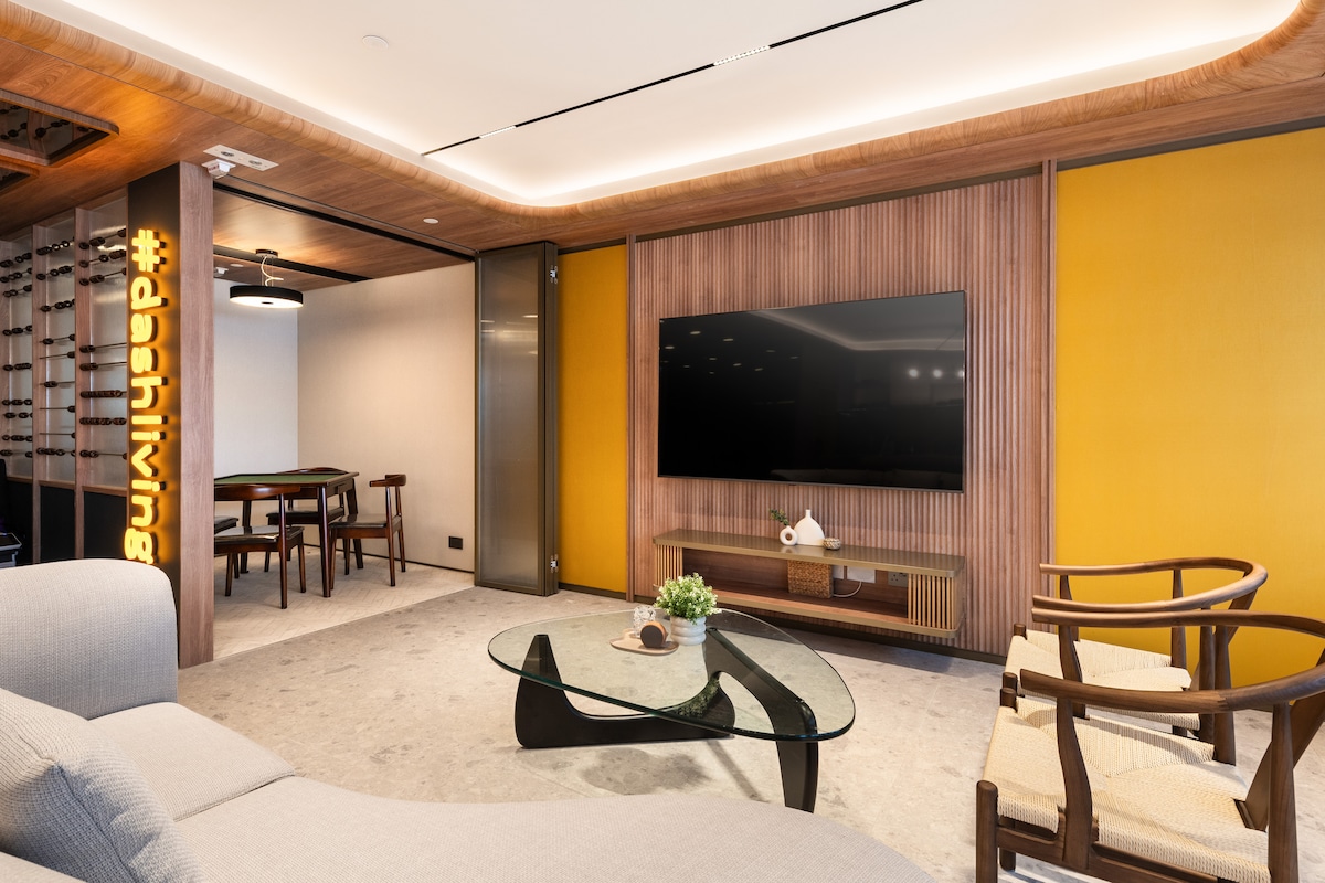A modern living area features warm wood accents and a sleek glass coffee table. A textured beige sofa is situated in front of a large television mounted on a wooden panel. Natural light enhances the space, while stylish chairs provide additional seating.