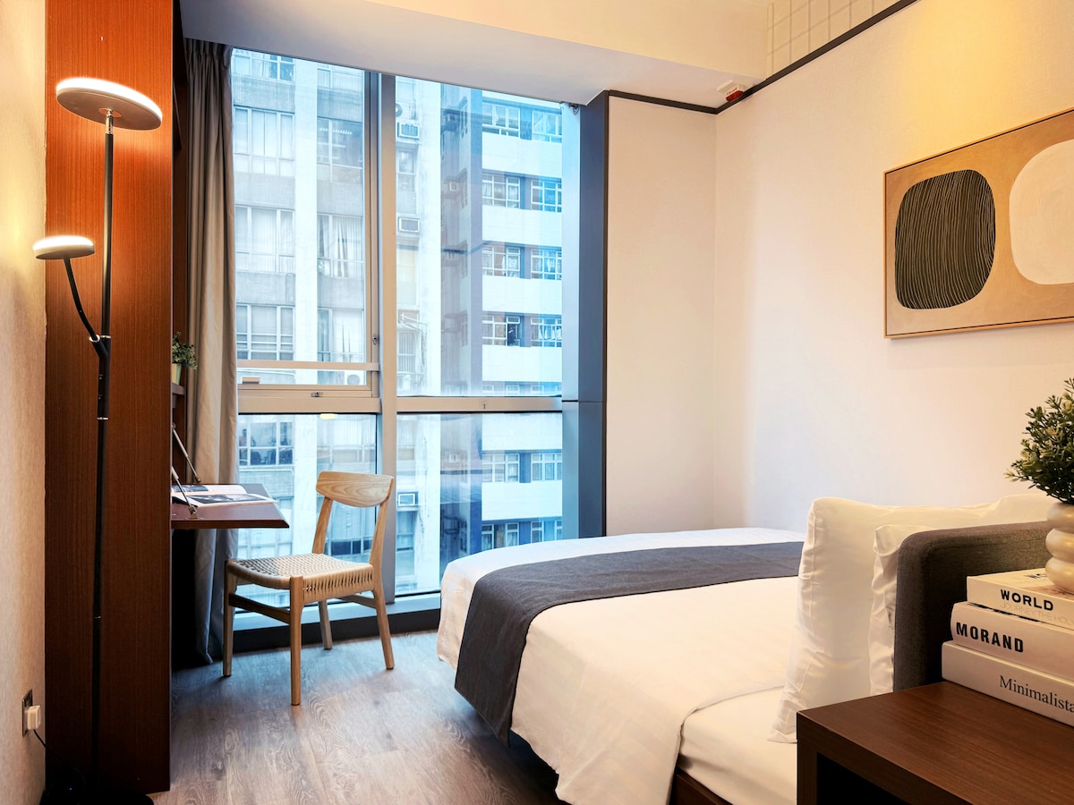 A bright room features a spacious bed with crisp white linens and a gray throw. A wooden chair and a small desk sit beside a large window that offers views of the surrounding buildings. Minimalist wall art complements the modern decor.