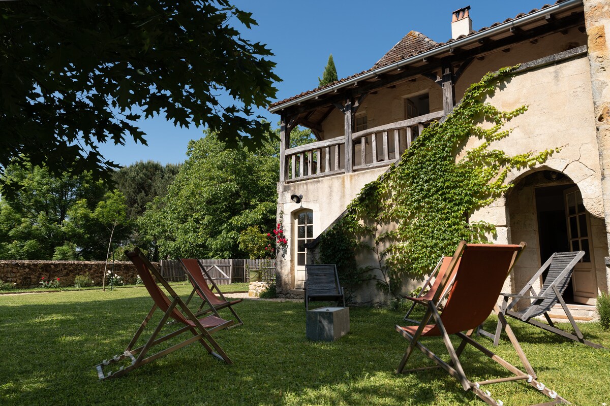 An inviting garden space surrounds the house, featuring a well-maintained lawn. Several lounge chairs are arranged for relaxation under the shade of trees. The upper balcony is accessible from the main floor, offering a vantage point of the natural surroundings.