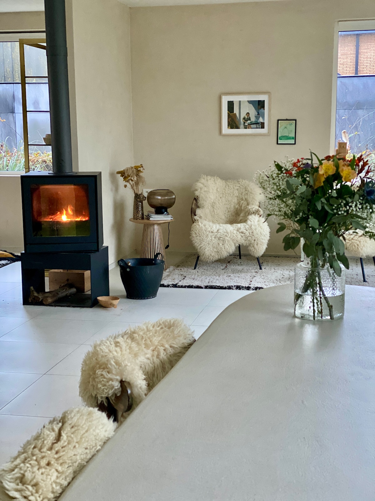 A warm living area features a modern wood-burning stove with a visible flame. Comfortable chairs adorned in soft, textured covers are positioned nearby. A vase filled with fresh flowers sits on a light-colored table, enhancing the space's inviting and natural atmosphere.