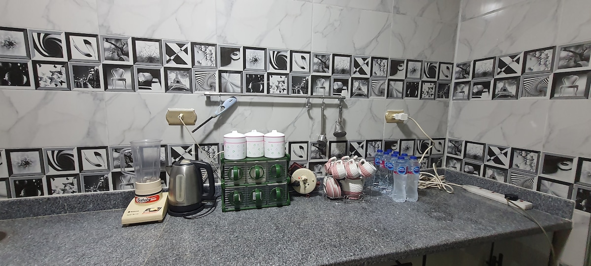 The kitchen features a granite countertop with various appliances including a blender, kettle, and a gas stove. A set of green glass containers is displayed alongside bottled water and a collection of cups. The patterned backsplash adds visual interest to the space.