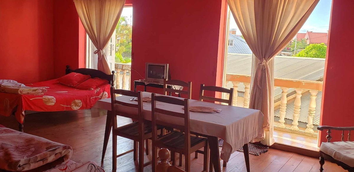 A bright room features large windows framed by flowing curtains, allowing natural light to fill the space. A dining table with several chairs is positioned nearby, while a bed with a colorful coverlet is visible. A small television is present, enhancing the comfort of the setting.
