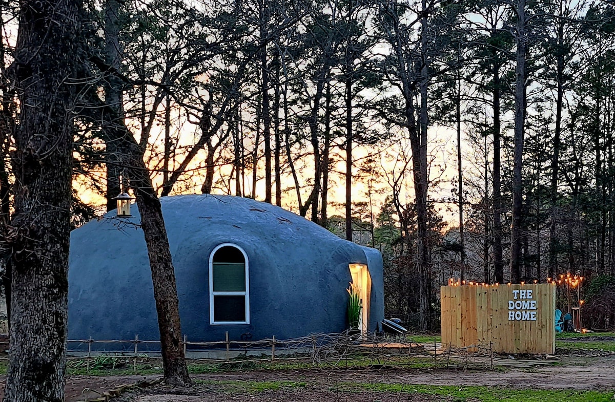 A unique dome-shaped home is nestled among tall trees, illuminated by the soft glow of sunset. A lighted sign reading 'THE DOME HOME' is positioned adjacent to the entrance, enhancing the serene ambiance of the natural surroundings.