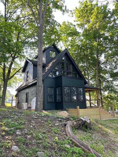 A modern, A-frame house is set in a wooded area, featuring tall windows that allow natural light to fill the space. A wooden deck with steps leads to the entrance, surrounded by lush greenery and a sloping hillside.