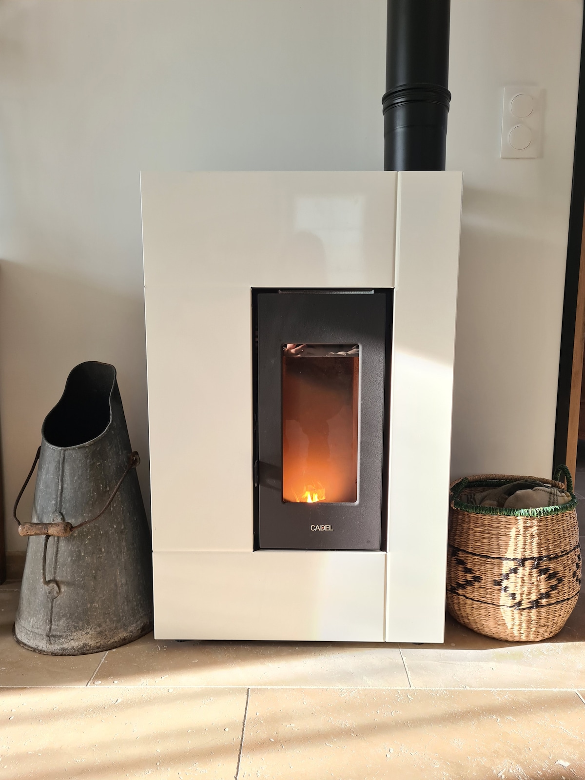 A sleek, modern stove is positioned against a light wall, featuring a viewing window displaying a flickering flame. Beside it, a metal container and a woven basket add a touch of natural texture to the setting.