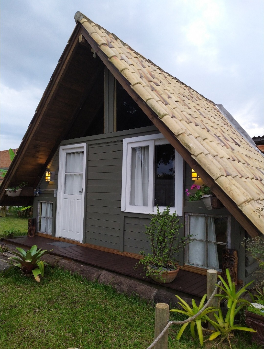 A charming cabin is showcased with a distinctive sloped roof and wooden accents. The exterior features multiple windows and plants strategically placed near the entrance, creating a harmonious connection with the surrounding greenery.