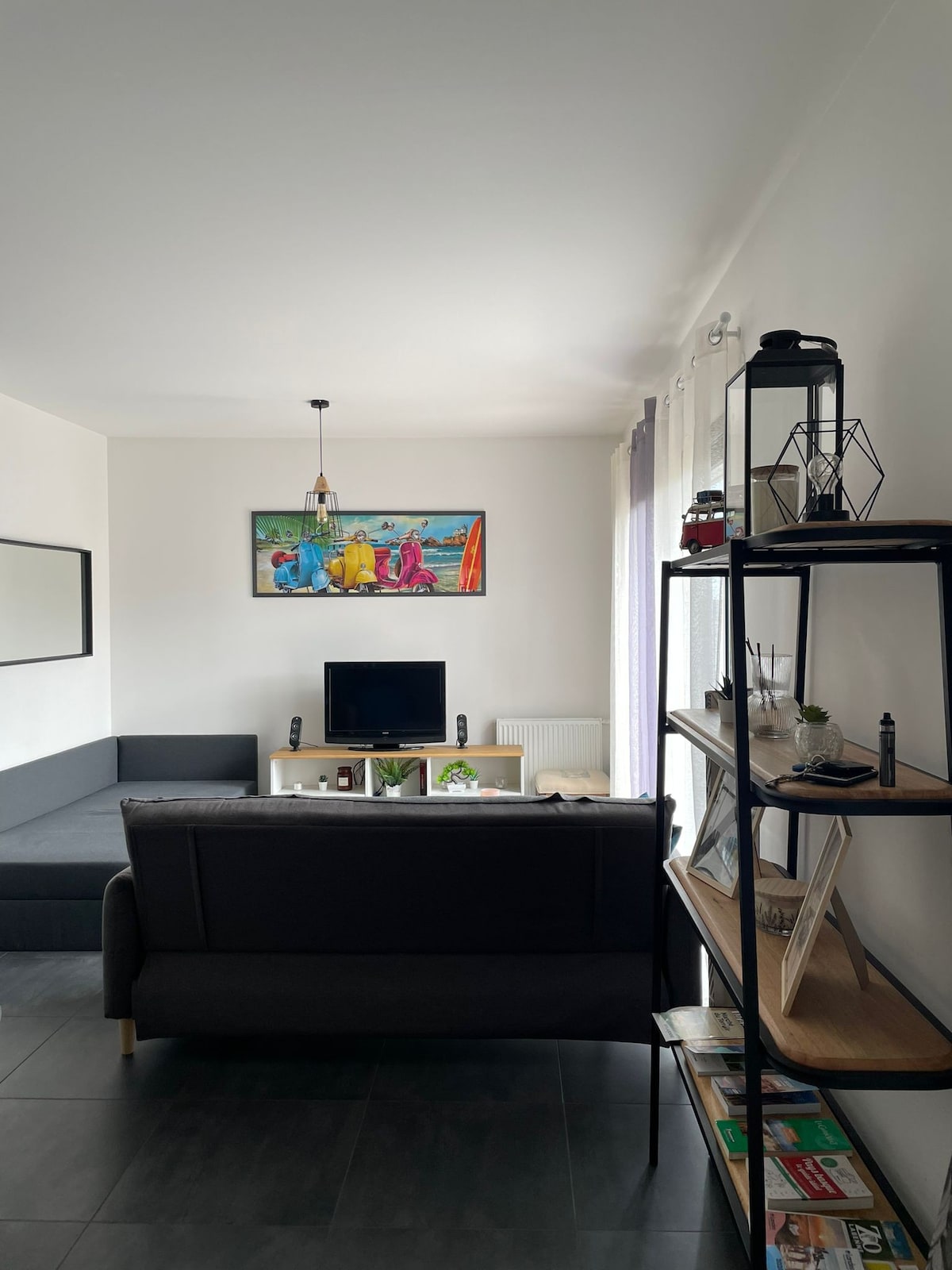 The living area features a dark sectional sofa and a light wooden surface with a television. A decorative painting hangs above the sofa, while a bookshelf displays magazines and plants. Natural light enters through window coverings, enhancing the space's open feel.