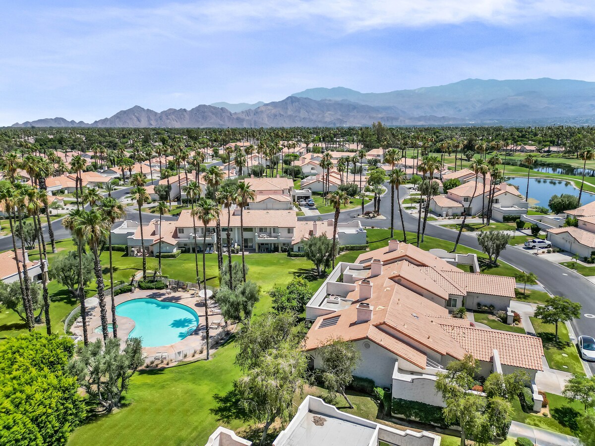 An aerial view showcases the well-maintained Desert Falls Country Club community, featuring neatly arranged villas surrounded by lush landscaping. A community pool is visible, along with palm trees adding a tropical ambiance. The mountainous backdrop enhances the serene desert setting.
