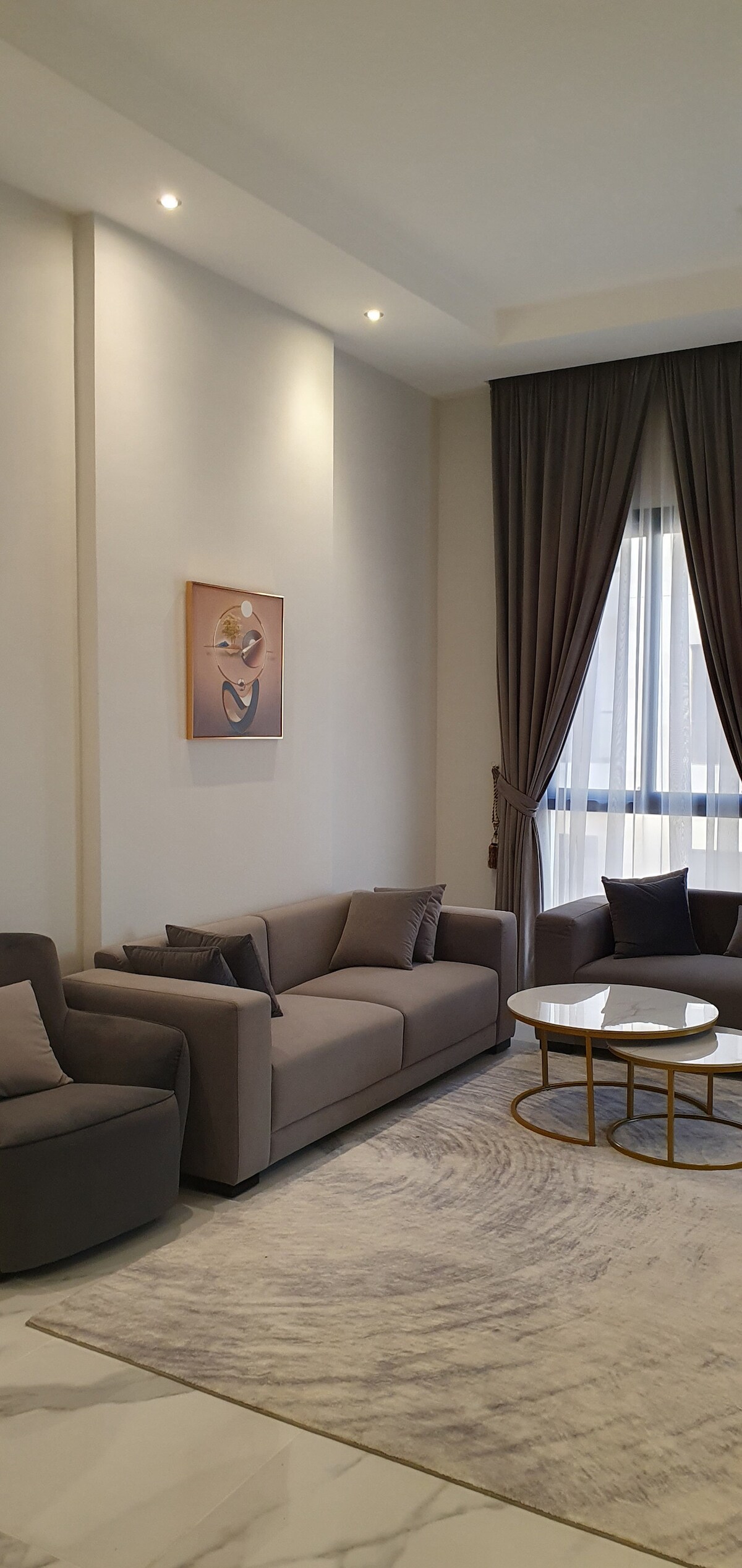 A spacious living area is highlighted by two soft gray sofas and an armchair, arranged for comfort. A round coffee table occupies the center, with decorative elements enhancing the design. Natural light filters through sheer curtains, illuminating the room's features.