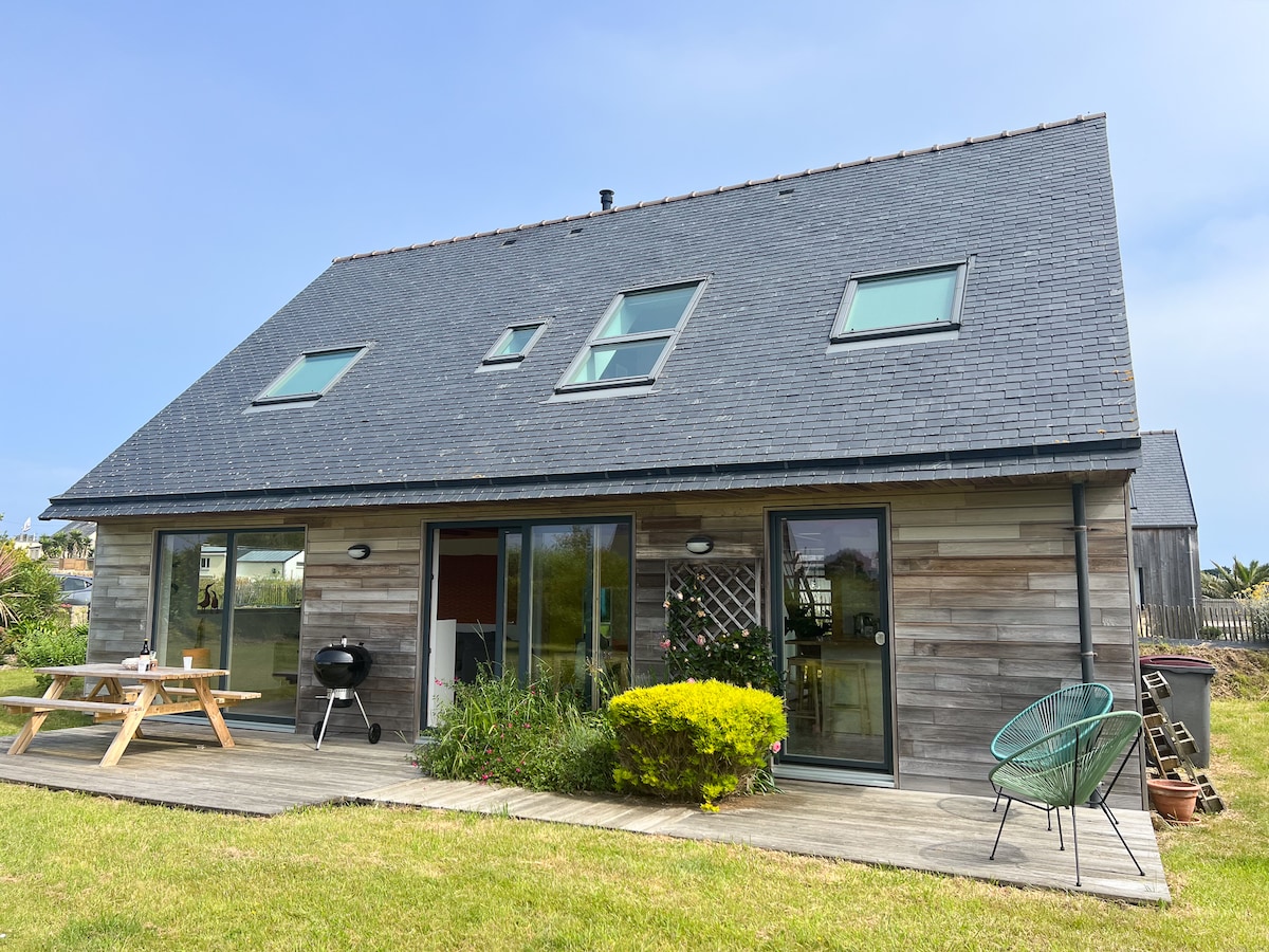 A two-story wooden house features a dark slate roof and large glass doors that open to a patio. A picnic table sits on the deck, accompanied by a small charcoal grill. Lush greenery surrounds the home, enhancing the outdoor space.