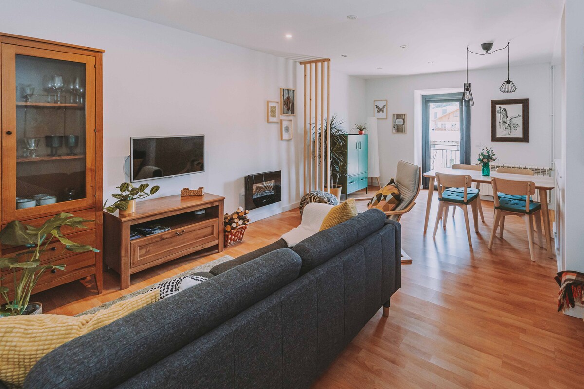 A well-lit living area features a comfortable gray sofa and wooden furniture, including a TV stand and a dining table. Plants are positioned throughout the space, and a balcony door provides natural light, enhancing the inviting atmosphere.