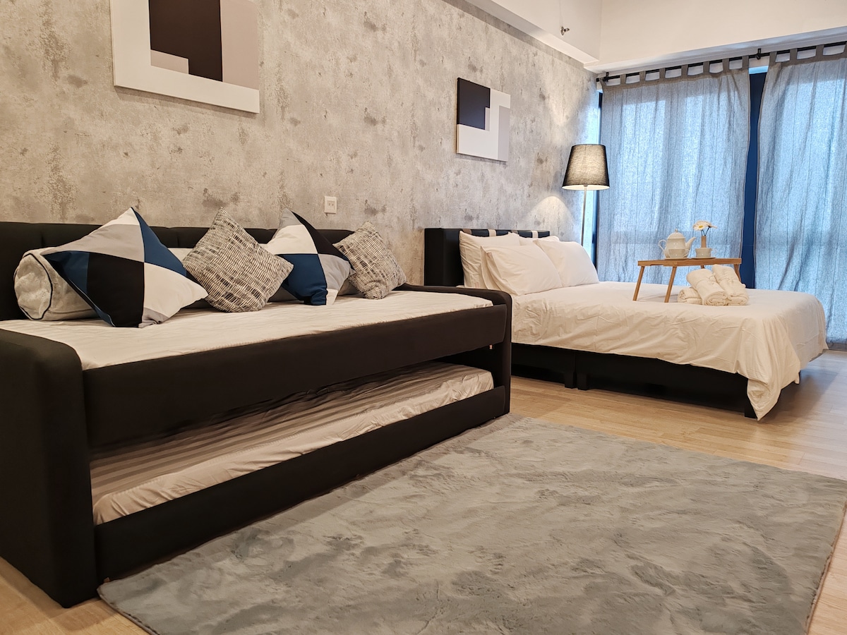 A well-designed studio apartment features a spacious bed and a comfortable sofa bed that can accommodate additional guests. Soft lighting from a nearby lamp creates a cozy ambiance, while large curtains frame the window, allowing natural light to enhance the room's inviting atmosphere.