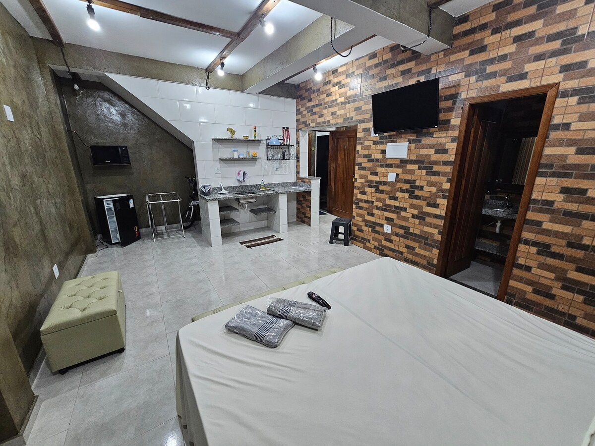 A spacious sleeping area features a neutral-toned bed with bedding neatly arranged. A kitchenette is visible with essential appliances such as a microwave and fridge. Textured walls in warm tones complement the modern decor, creating a welcoming ambiance.