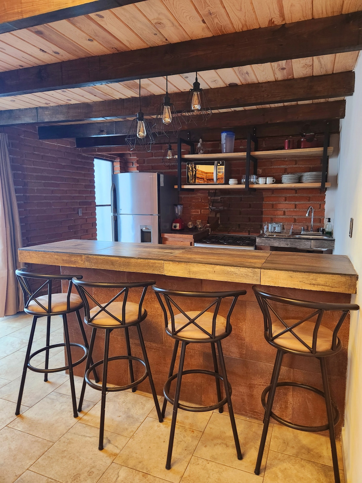The kitchen area features a wooden bar with four high-backed stools. Brick walls contrast with modern appliances, including a refrigerator and stove. Exposed wooden beams run across the ceiling, with pendant lighting adding a functional touch. Shelves display various dishes and kitchen essentials.