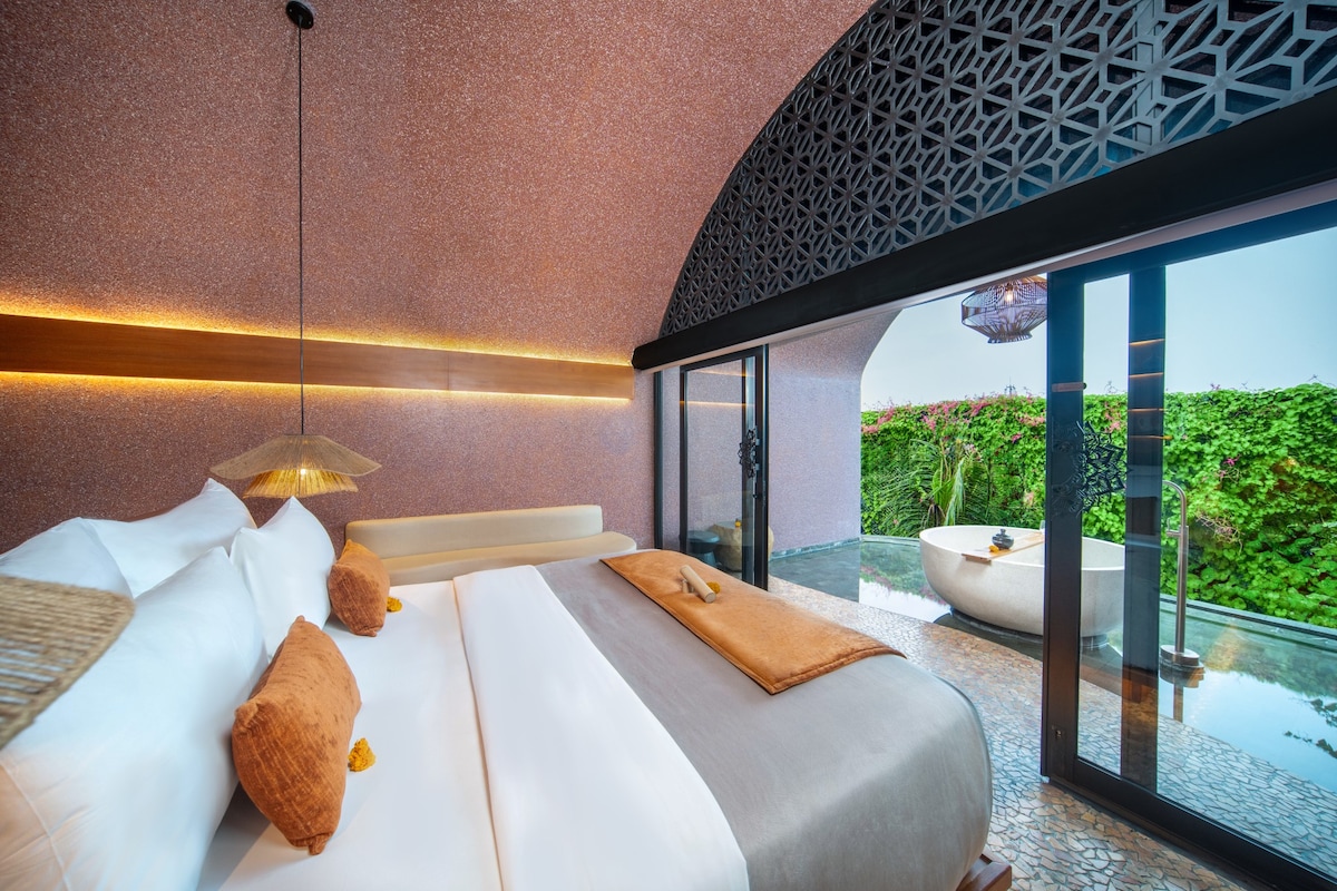 A spacious bedroom is featured with an elegant bed adorned with soft pillows and an earthy-toned blanket. Large glass doors provide access to a private outdoor area, framed by lush greenery. Subtle lighting enhances the warm color palette, contributing to a tranquil atmosphere.