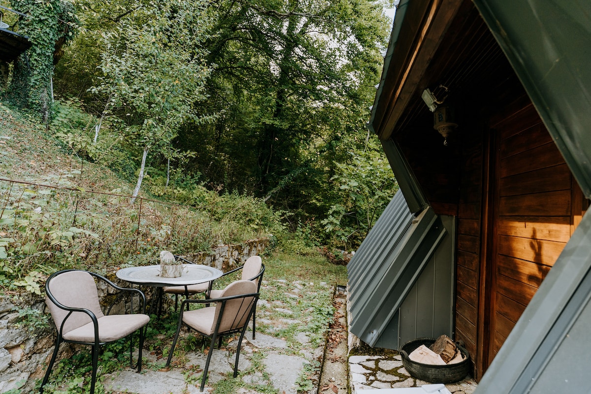 A seating area is set outside the lodge, featuring a round table surrounded by four elegant chairs. The natural stone pathway leads to the entrance, framed by lush greenery that enhances the tranquil setting.