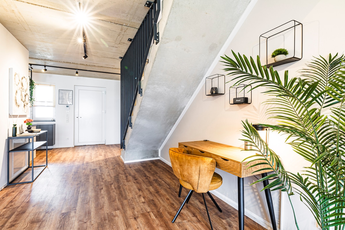 The entry area features a wooden table positioned near a large green plant. A chair with a warm hue is placed at the table, while decorative wall-mounted planters add a touch of greenery. Light from an overhead fixture illuminates the space, enhancing its inviting appearance.
