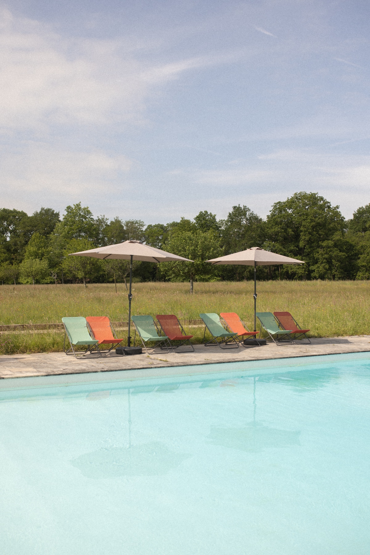 A serene pool area is visible, featuring crystal-clear water and several lounge chairs with colorful cushions under large umbrellas. Green trees and an expansive grassy field surround the space, creating a tranquil outdoor atmosphere.