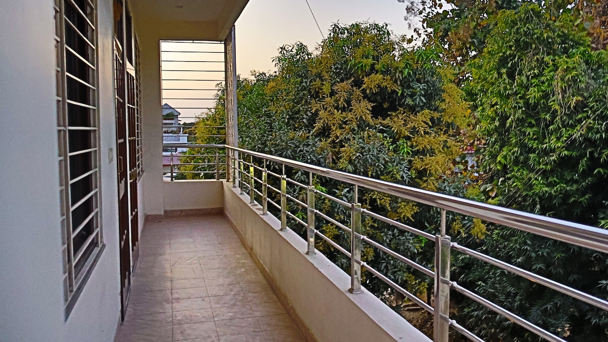 A spacious balcony is shown, bordered by a sleek metal railing. Lush greenery is visible in the surrounding area, adding a natural touch. The flooring is tiled, and the early morning sun casts a gentle light, enhancing the serene atmosphere.