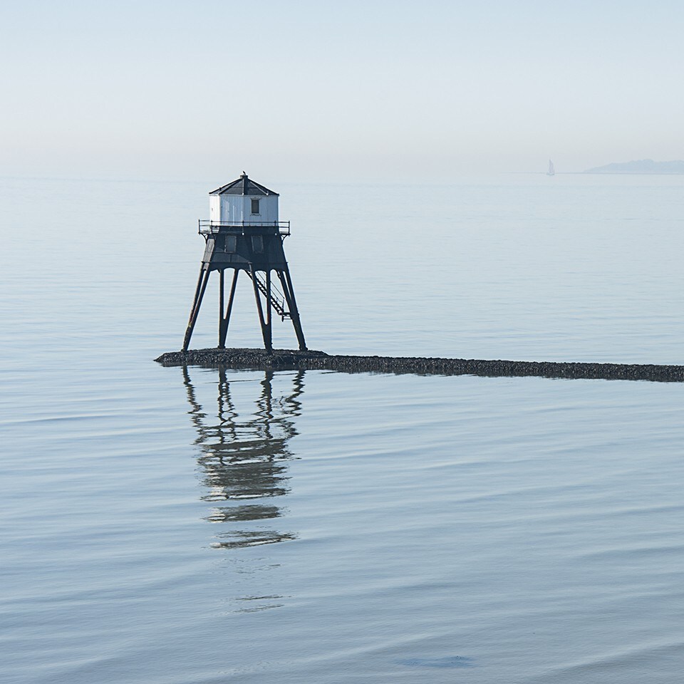 A solitary lighthouse stands on stilts above calm waters, creating a serene focal point. The stillness is reflected in the smooth surface of the sea, enhancing the peaceful atmosphere. A faint outline of land can be seen in the distance, adding depth to the tranquil scene.