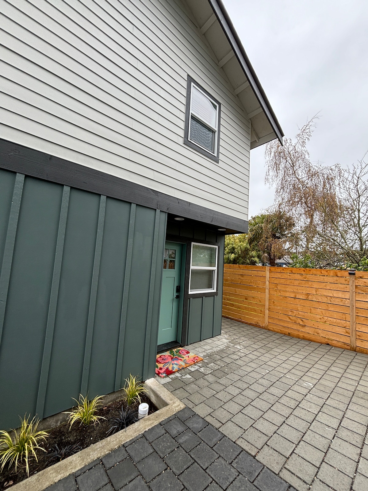 The exterior of a two-level cottage is presented, showcasing a combination of textured siding in muted tones. A welcoming entrance features a vibrant door, with a small porch area adorned with colorful doormats. Neat landscaping and a wooden privacy fence frame the yard.