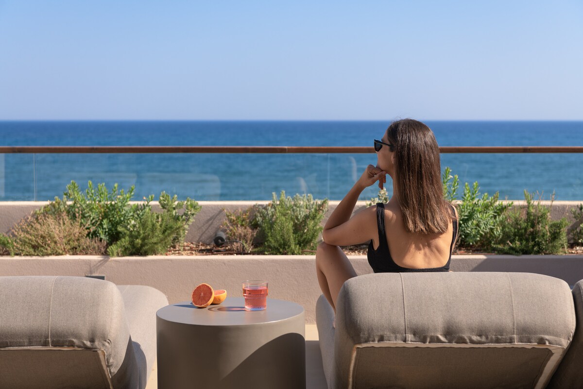 A serene outdoor seating area features two soft armchairs positioned towards the expansive view of the sea. A small table with a refreshing drink, accompanied by citrus slices, rests between the chairs. Verdant plants enhance the coastal ambiance under a clear blue sky.