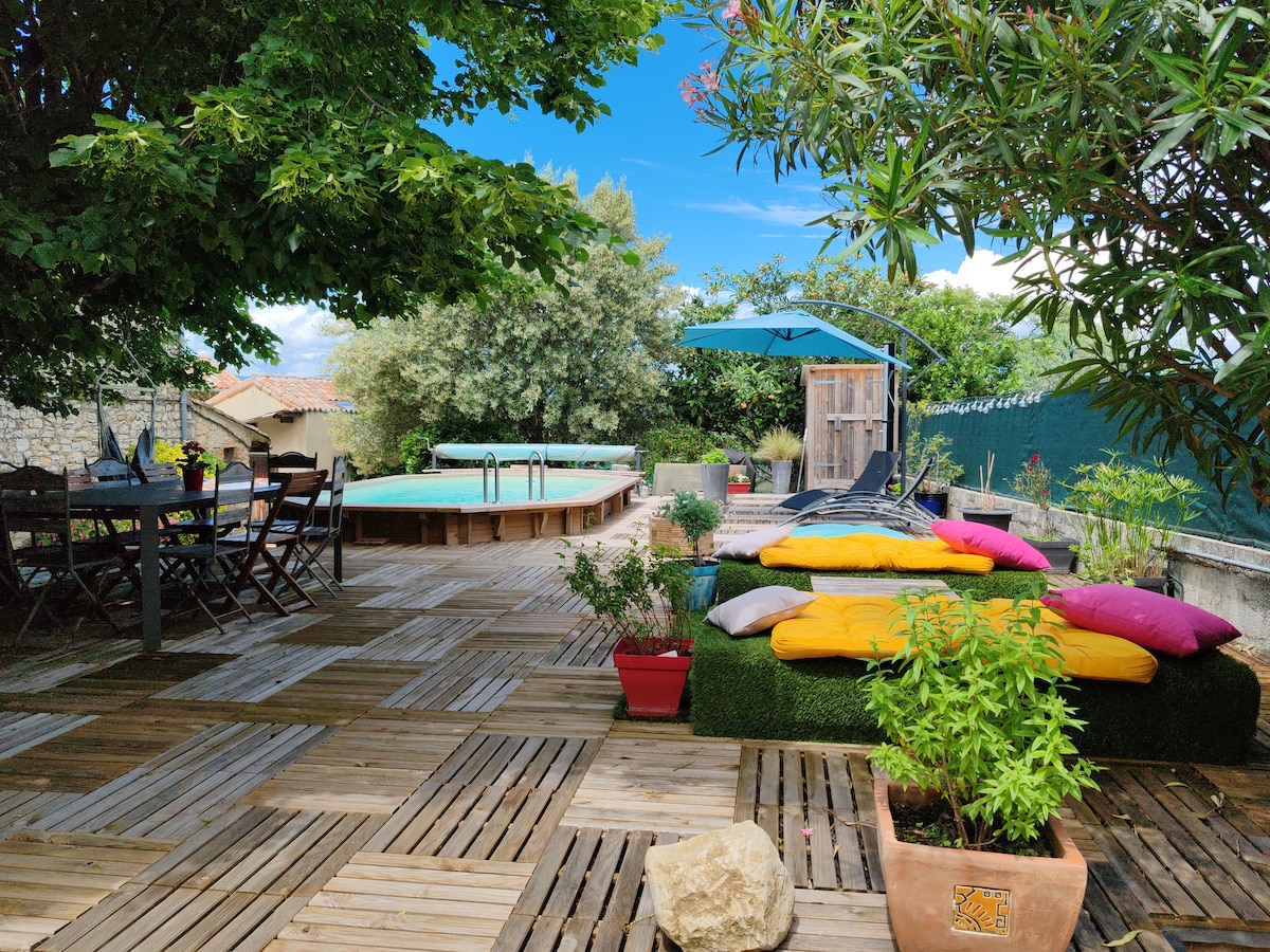 A spacious outdoor terrace features a swimming pool and wooden decking, surrounded by greenery. Lounge chairs are arranged near the pool, alongside several colorful cushions on a grassy area. A shaded seating area includes a table with chairs, inviting relaxation in a tranquil setting.