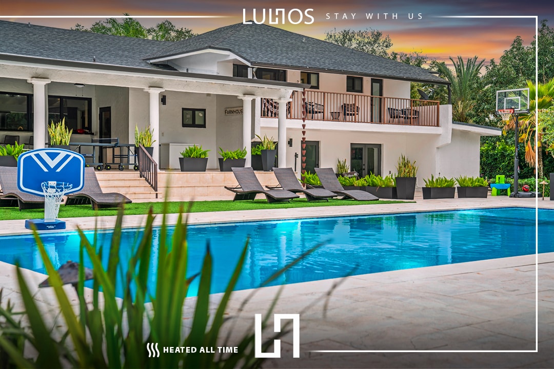 The outdoor area features a spacious heated pool surrounded by sun loungers and lush greenery. The two-story villa stands in the background, showcasing a balcony with seating. A basketball hoop is positioned nearby, enhancing the recreational atmosphere.