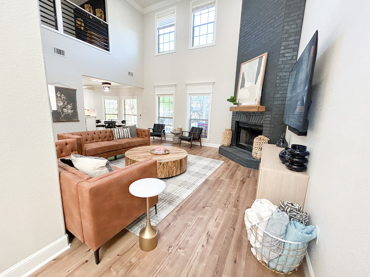 A spacious living area features a large sectional sofa arranged around a round wooden coffee table. Natural light fills the room through tall windows. A fireplace adds a cozy focal point, complemented by decorative elements on shelves and a basket holding soft throws.