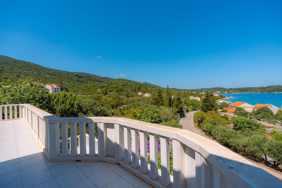 A spacious terrace is shown, featuring elegant railings and tiles that reflect sunlight. The view captures lush green hills, vibrant coastal homes with terracotta roofs, and the serene blue waters of the Adriatic Sea under a clear sky.