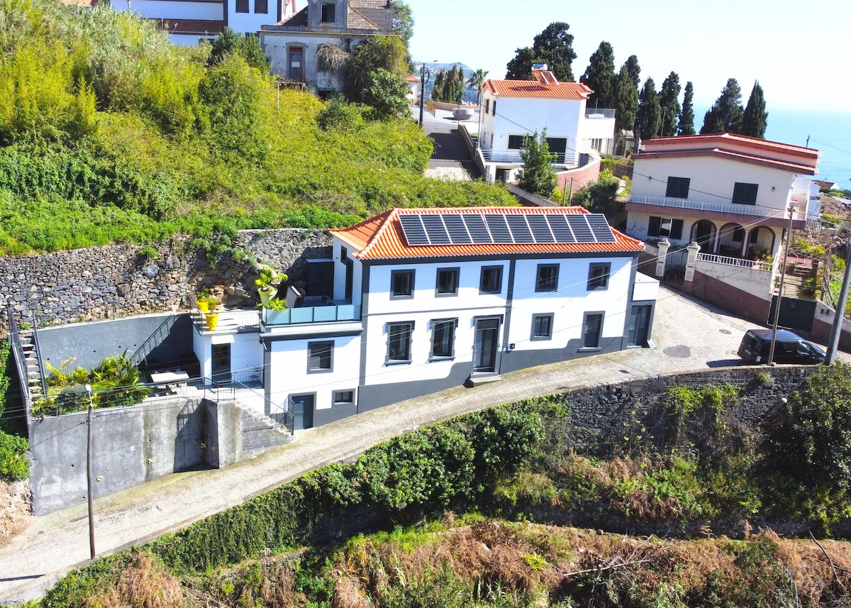 An aerial view of a spacious villa is displayed, showcasing a well-maintained exterior painted in white with black accents. Solar panels are positioned on the roof. Verdant hills and nearby homes are visible in the background, with the shimmering sea in the distance.
