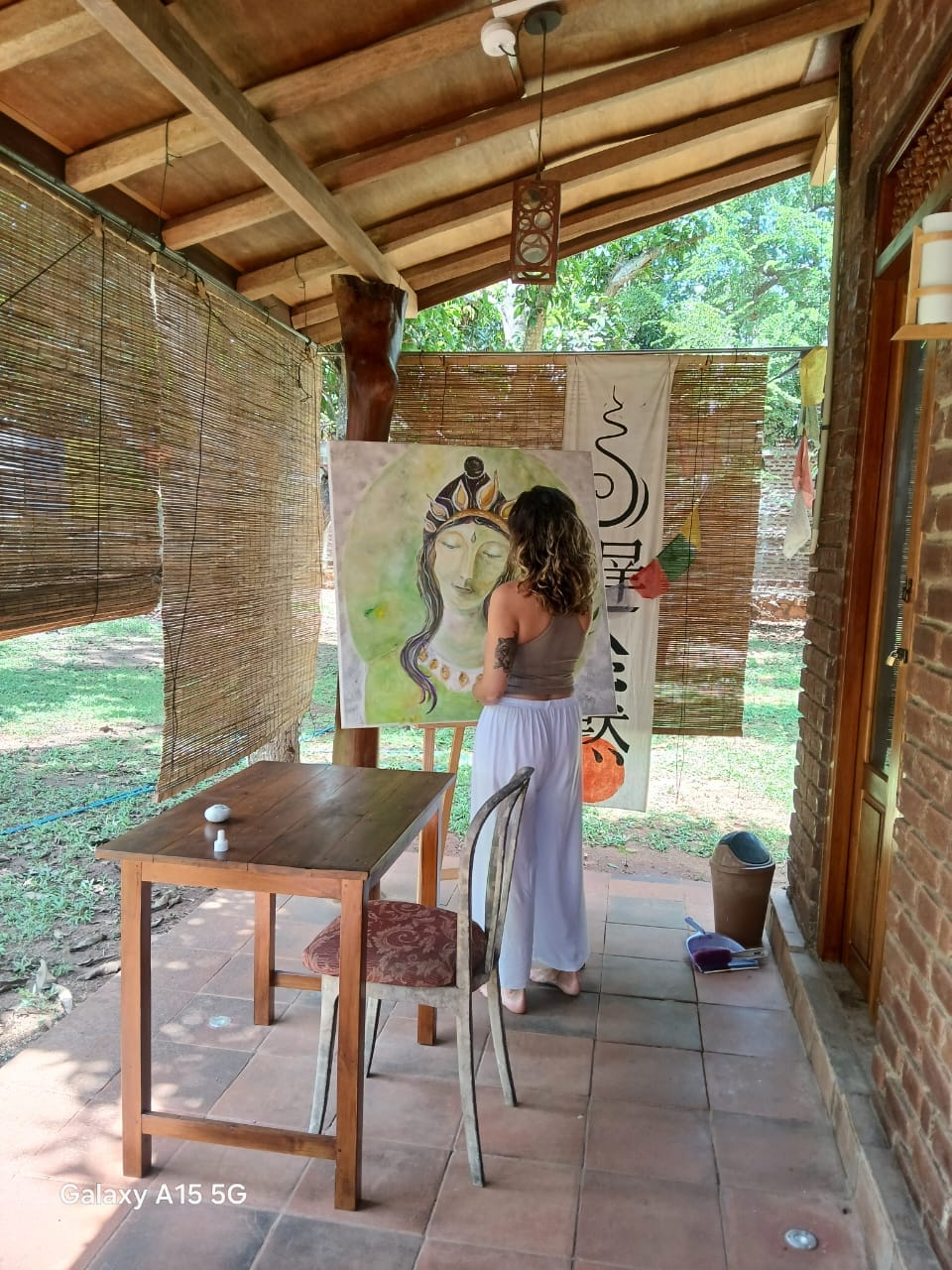 An artist is seen painting on a canvas within a rustic studio space. Natural light fills the area, complemented by wooden beams and bamboo blinds. A small table with a single chair is positioned nearby, and the rich greenery of the garden is visible in the background.