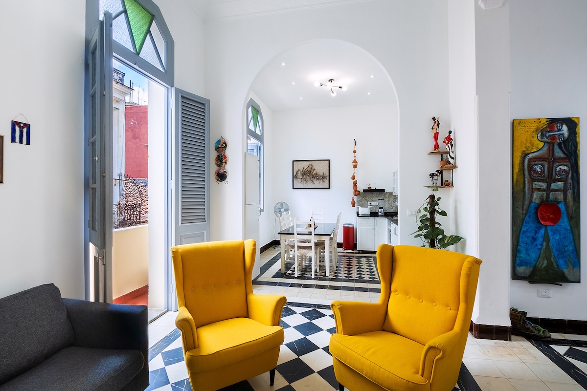 A bright and airy living area features two yellow armchairs with a black and white checkered floor. A brown sofa is positioned nearby, while an open archway leads to a kitchen and dining space. Colorful artwork and decorative elements add character to the room.
