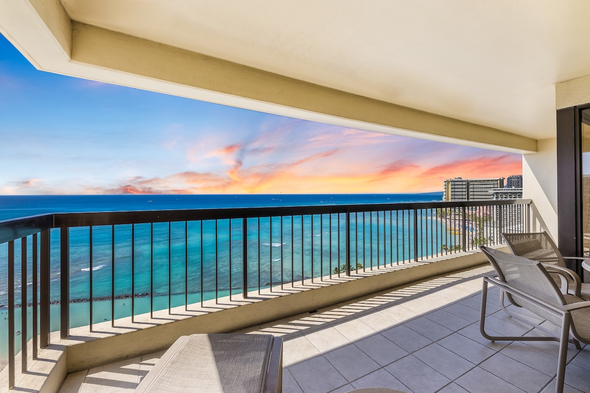 An expansive lanai offers stunning views of the ocean, with a backdrop of vibrant sky colors at sunset. Comfortable outdoor seating is provided, inviting relaxation while enjoying the scenery. The balcony railing allows for unobstructed sightlines of the water's edge.