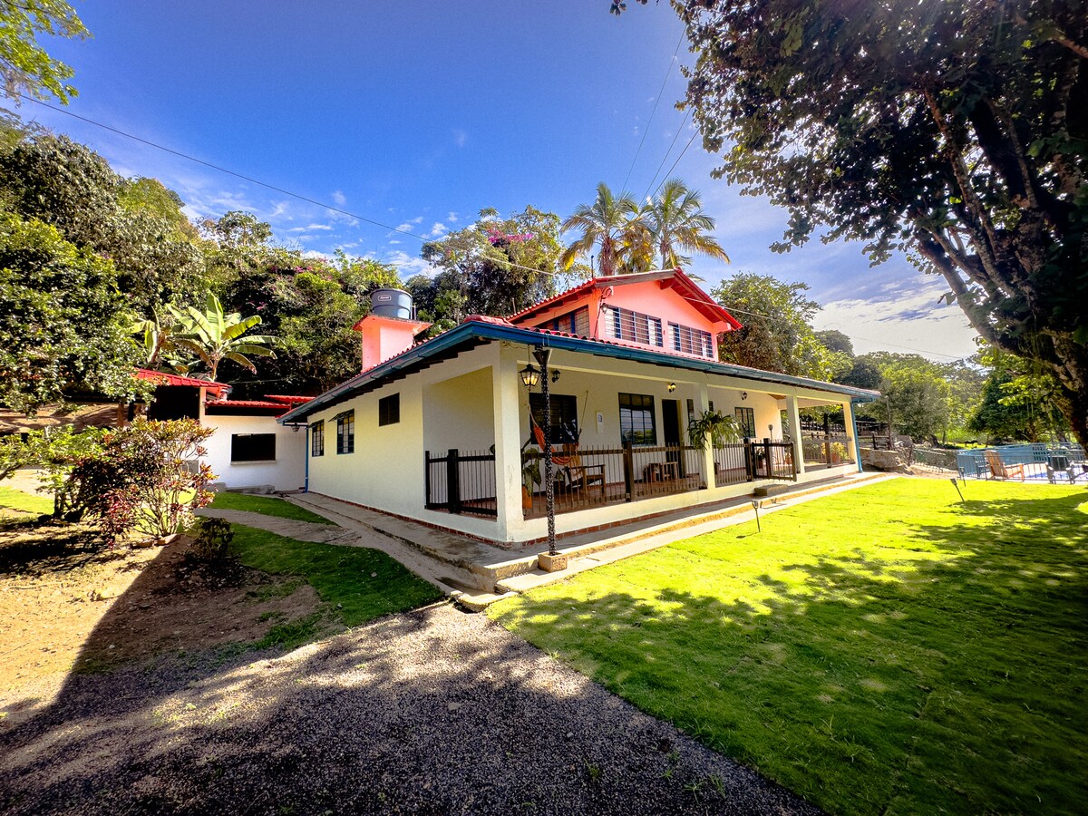 A two-story house is set amidst lush greenery, featuring a red roof and a combination of light and dark exterior walls. A spacious front porch with seating is visible, surrounded by well-maintained grass and trees. A pool area can be seen in the background.