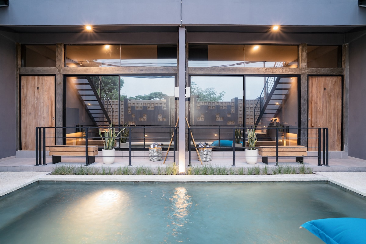 A tranquil pool reflects the evening light, framed by two modern loft units with large glass doors. Wooden beams enhance the contemporary design, while potted greenery and outdoor seating create a welcoming atmosphere. A staircase is visible in the background, leading to upper-level units.