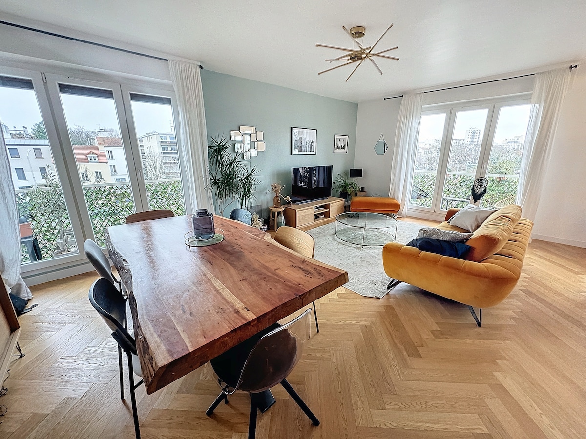 A spacious living area features large windows showcasing city views. A wooden dining table is surrounded by black chairs, while a comfortable orange sofa and glass coffee table add a modern touch. Natural light illuminates the neutral walls, enhancing the room's inviting atmosphere.