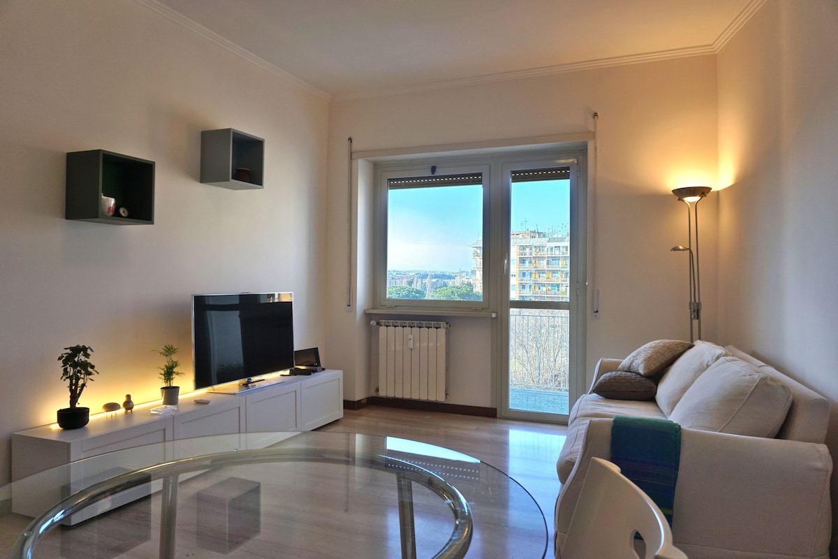 A well-lit living area features a comfortable sofa, a modern entertainment unit, and a television. Large windows provide natural light and a view of the surrounding area. A small plant adds a touch of greenery, while minimalistic shelves display decorative items.