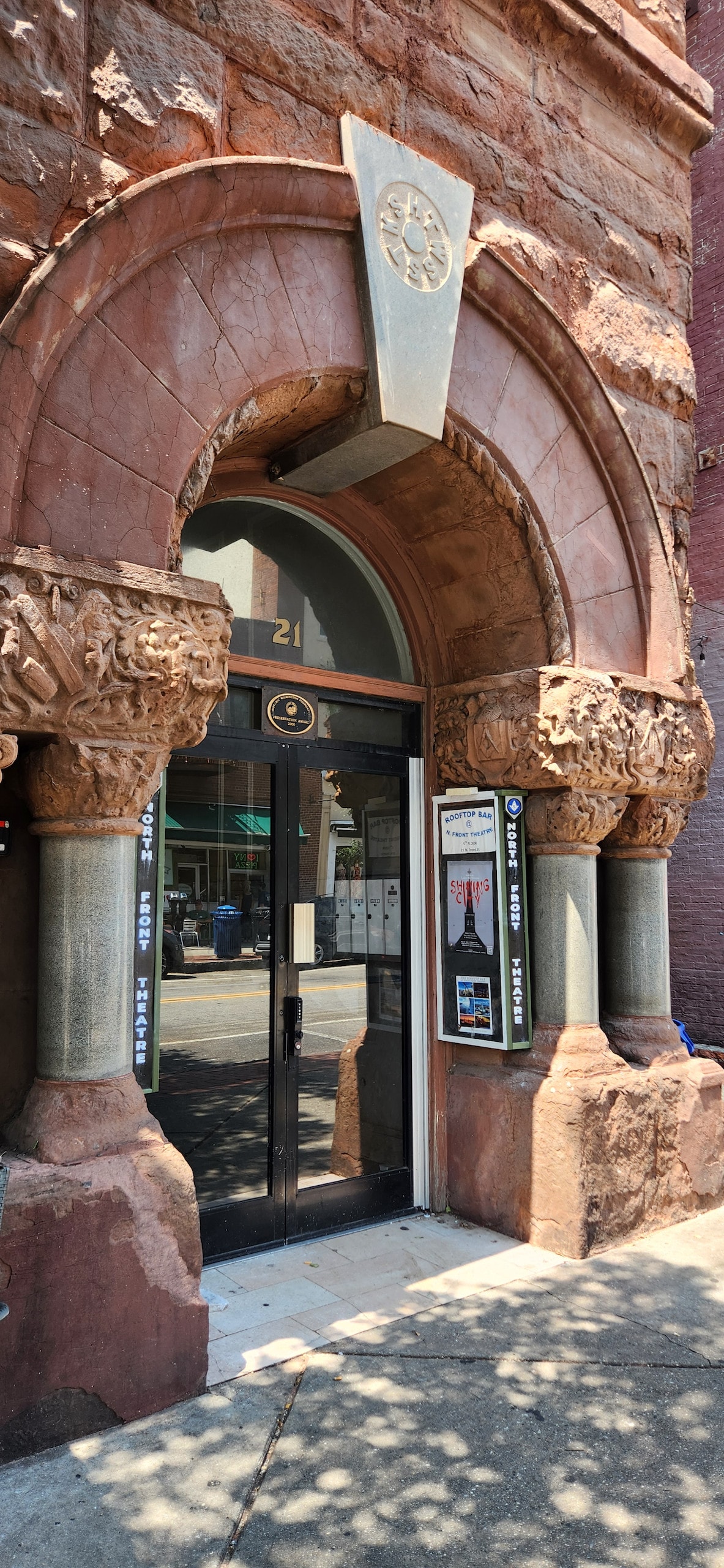 The image captures the entrance of a historic building, featuring a detailed archway adorned with intricate carvings. The door is framed with dark glass, and nearby, informational posters are displayed. Natural light creates a dappled effect on the sidewalk below.