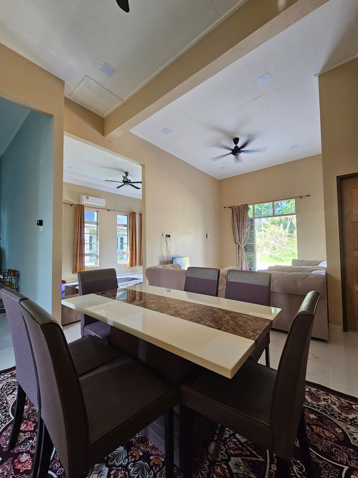 A dining area is showcased with a rectangular table surrounded by four upholstered chairs in neutral tones. The space is illuminated by natural light filtering through a window. In the background, a living area is visible, featuring a comfortable sofa and ceiling fan.