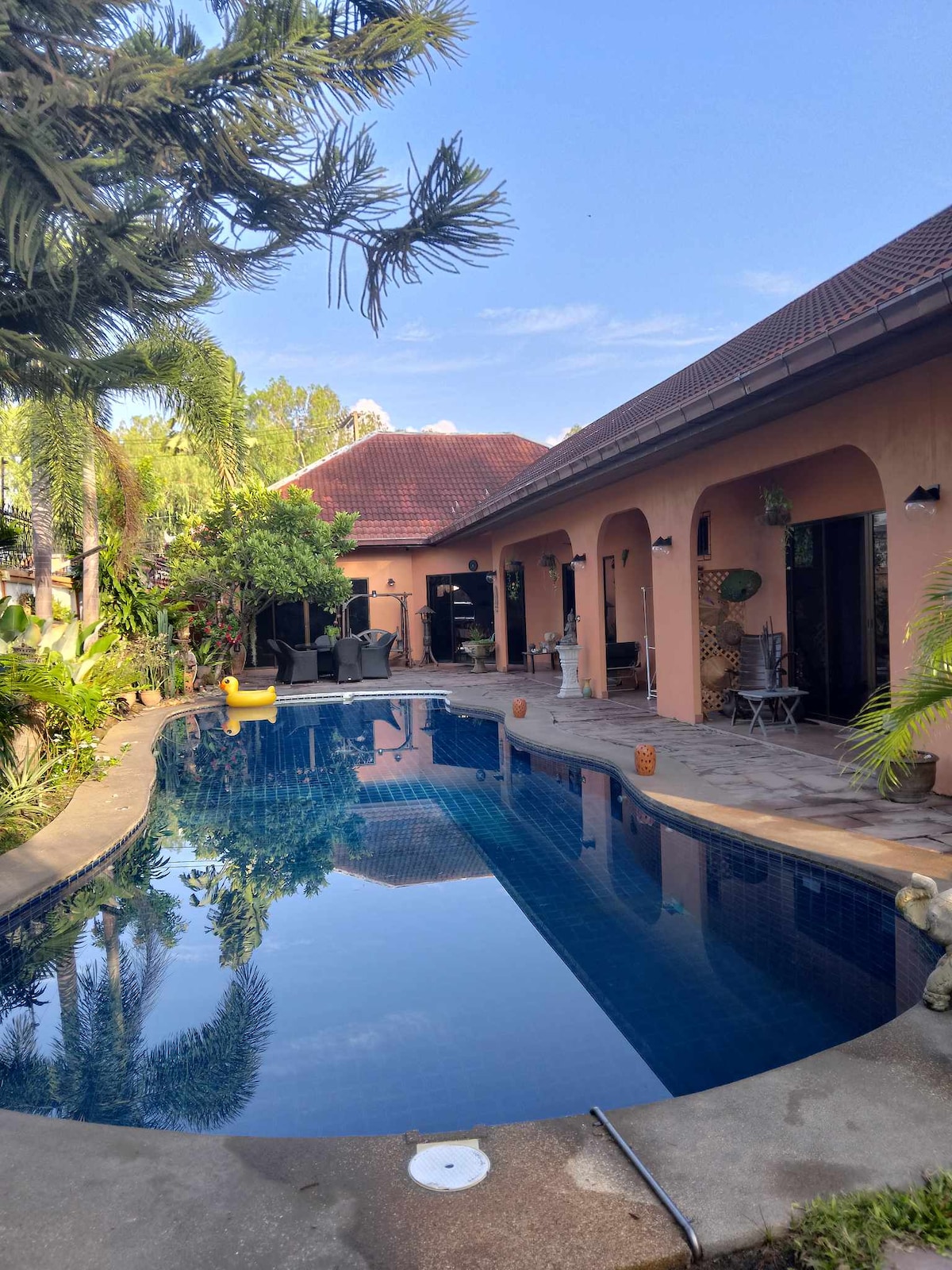 A serene outdoor area surrounds a reflective swimming pool. Lush greenery frames the space, with a playful duck float resting on the water. The building features an array of arches, and sun loungers are positioned nearby, inviting relaxation.